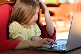 Young girl sitting in her mother's lap using a laptop to symbolize how online testing requires keyboard and mouse skills.